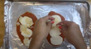 Hands preparing chicken parmesan with tomato sauce and cheese on foil-lined baking sheet.